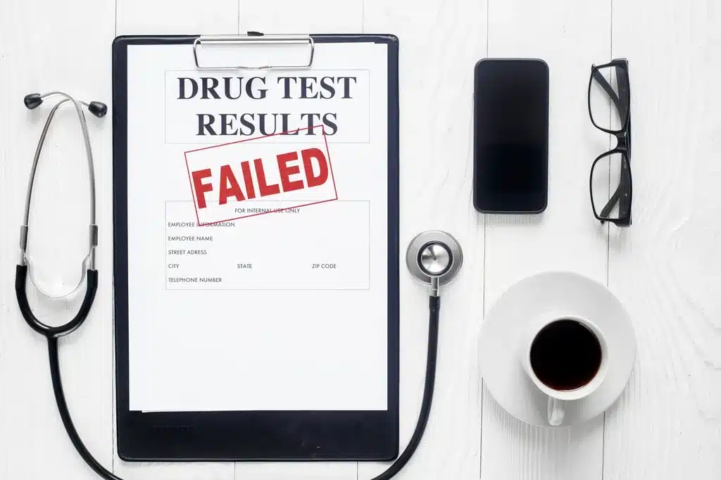 A failed drug test results document, stethoscope, phone, eyeglasses, and coffee on a table. 